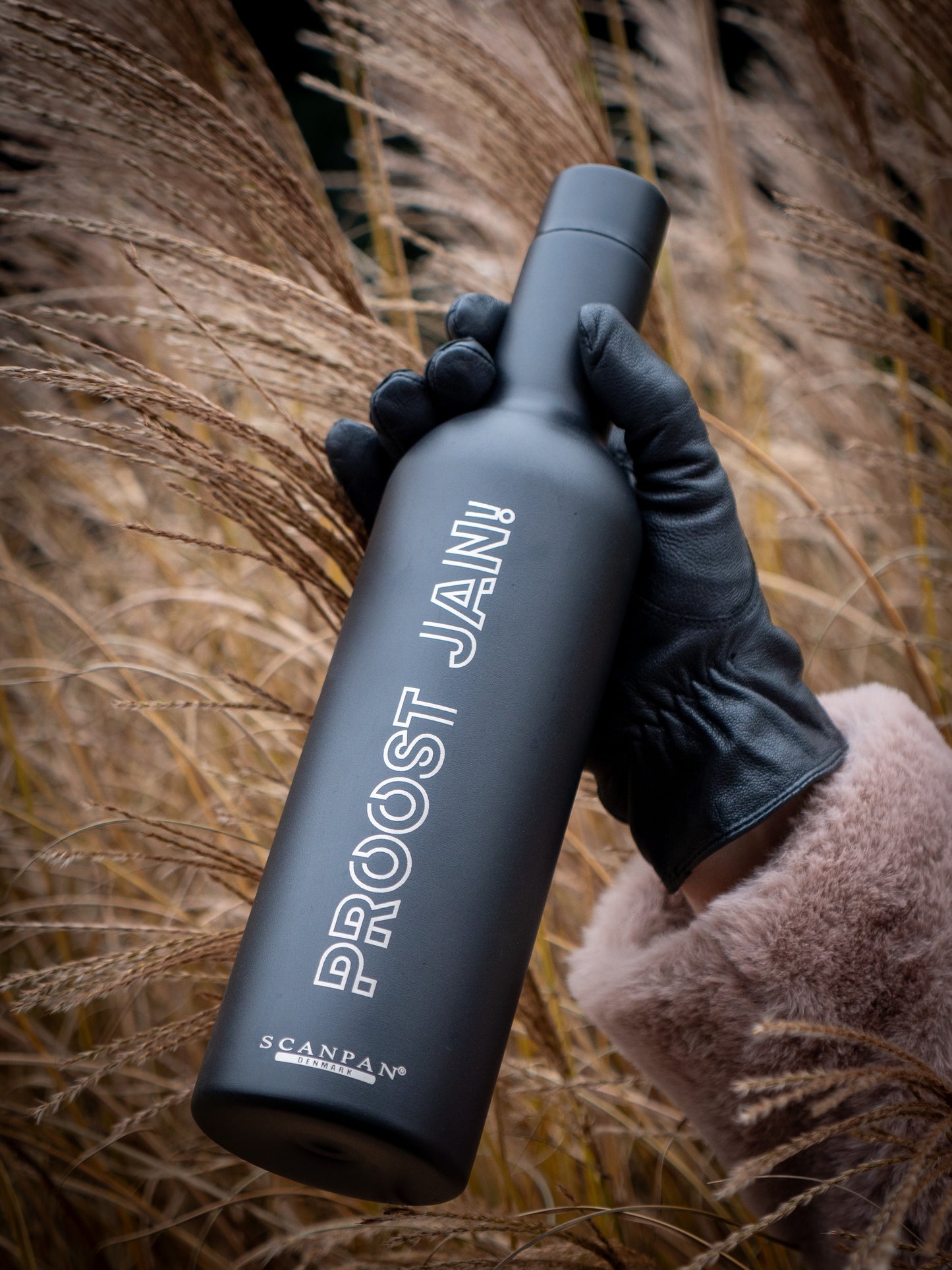 Thermosfles met proost jan er op in een leren hand met op de achtergrond riet
